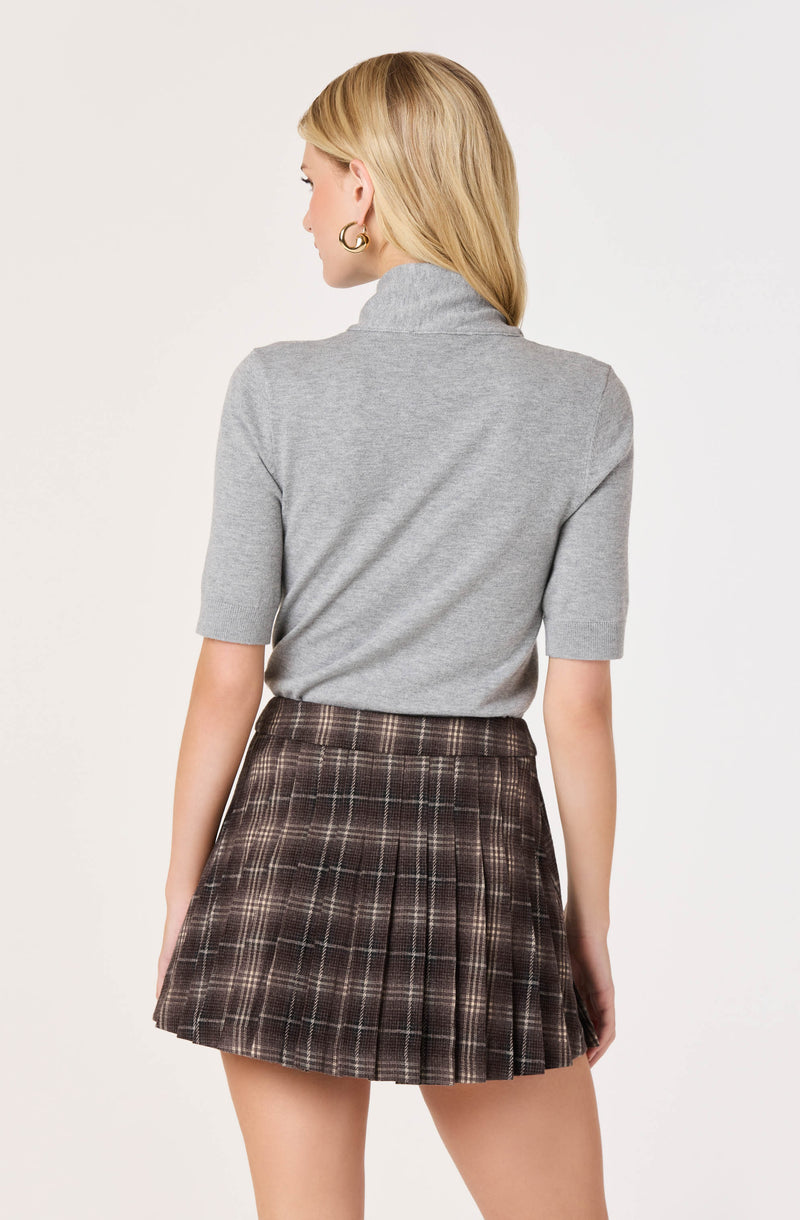 Woman wearing a light gray turtleneck sweater and a brown plaid pleated skirt.
