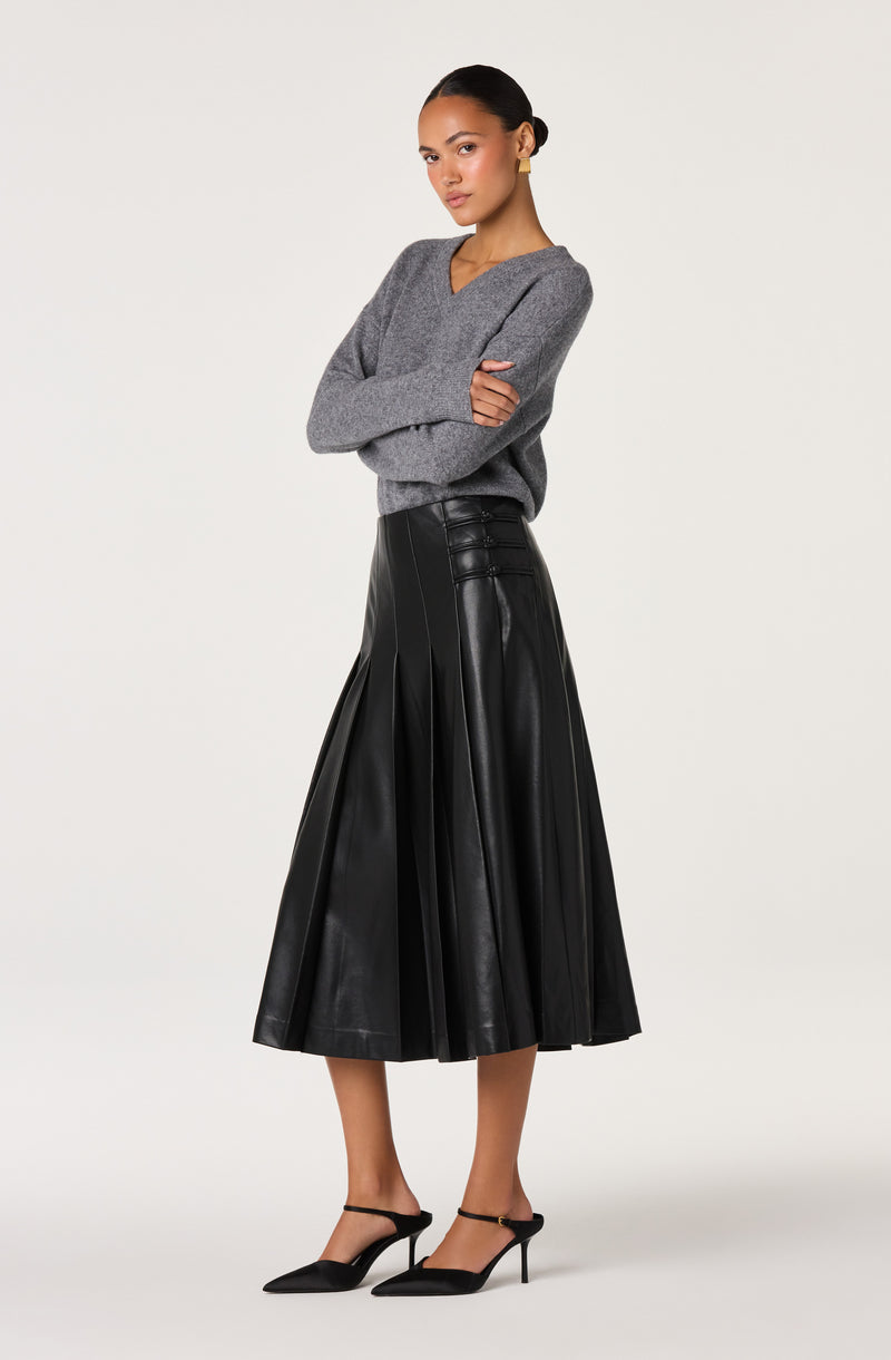 Woman wearing a gray V-neck sweater, black leather pleated midi skirt, and black high heels.