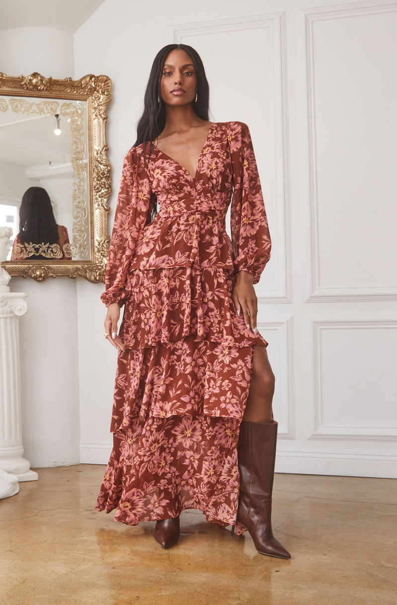 The dress is a long, flowy, floral print gown in shades of pink and burgundy. It features a V-neckline, long puffed sleeves, a fitted waist, and layered ruffles throughout the skirt. The dress has a high slit on one side, revealing a pair of knee-high brown leather boots.