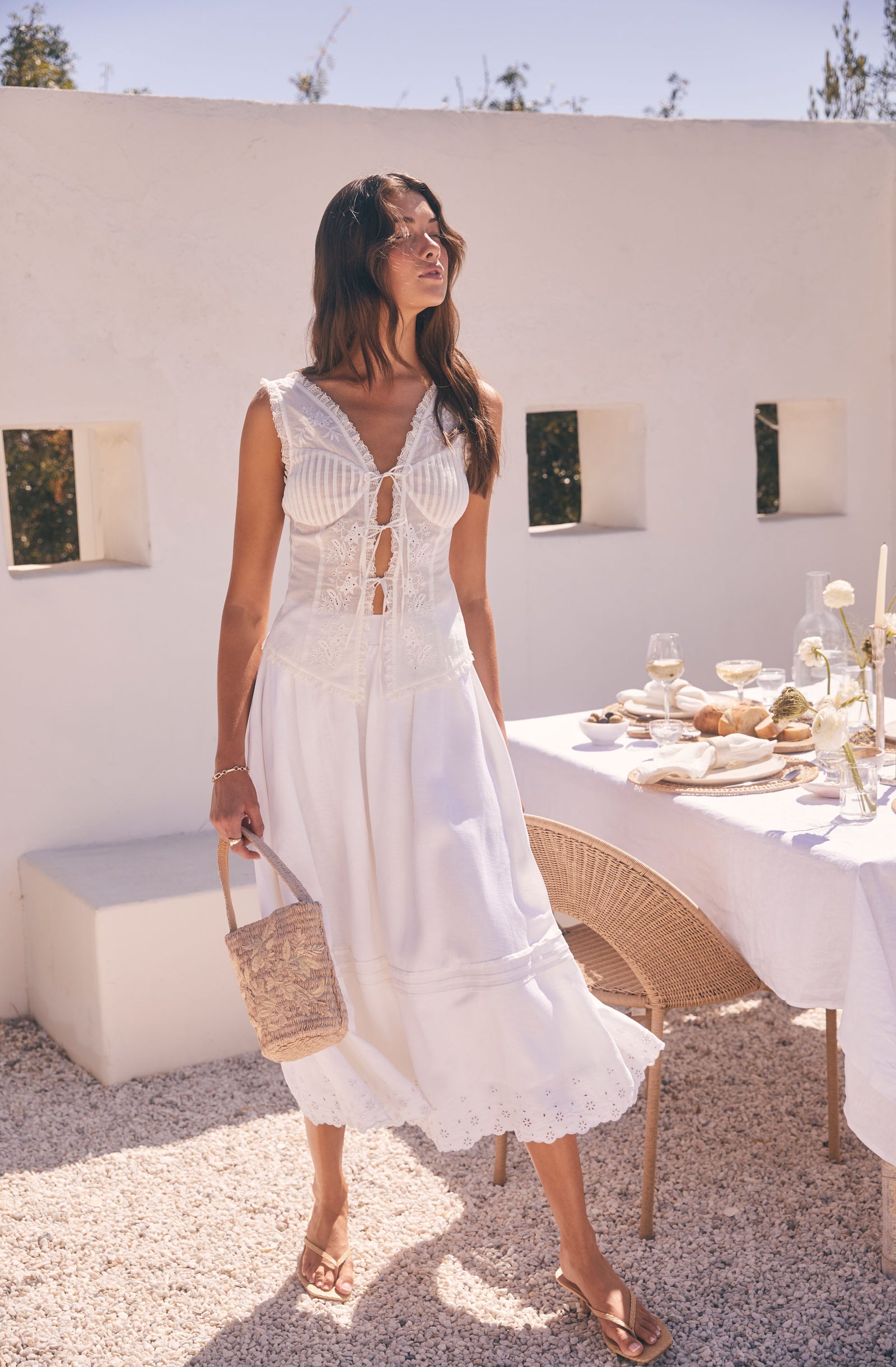 Woman in white sleeveless, embroidered dress holding a straw handbag outdoors.