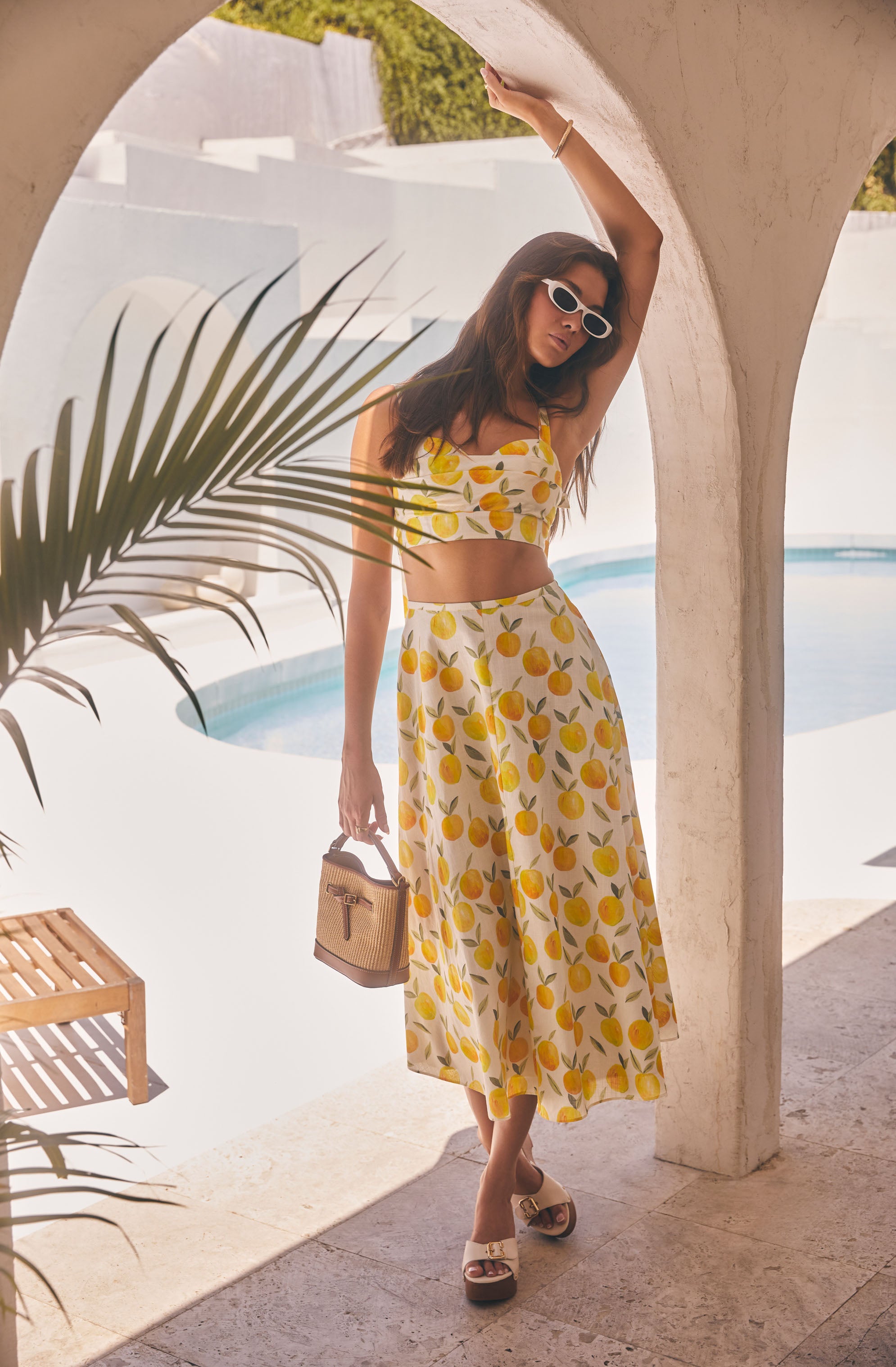 Woman in yellow lemon print matching crop top and maxi skirt, white sunglasses, holding a small woven bag, standing under an arch near a pool.
