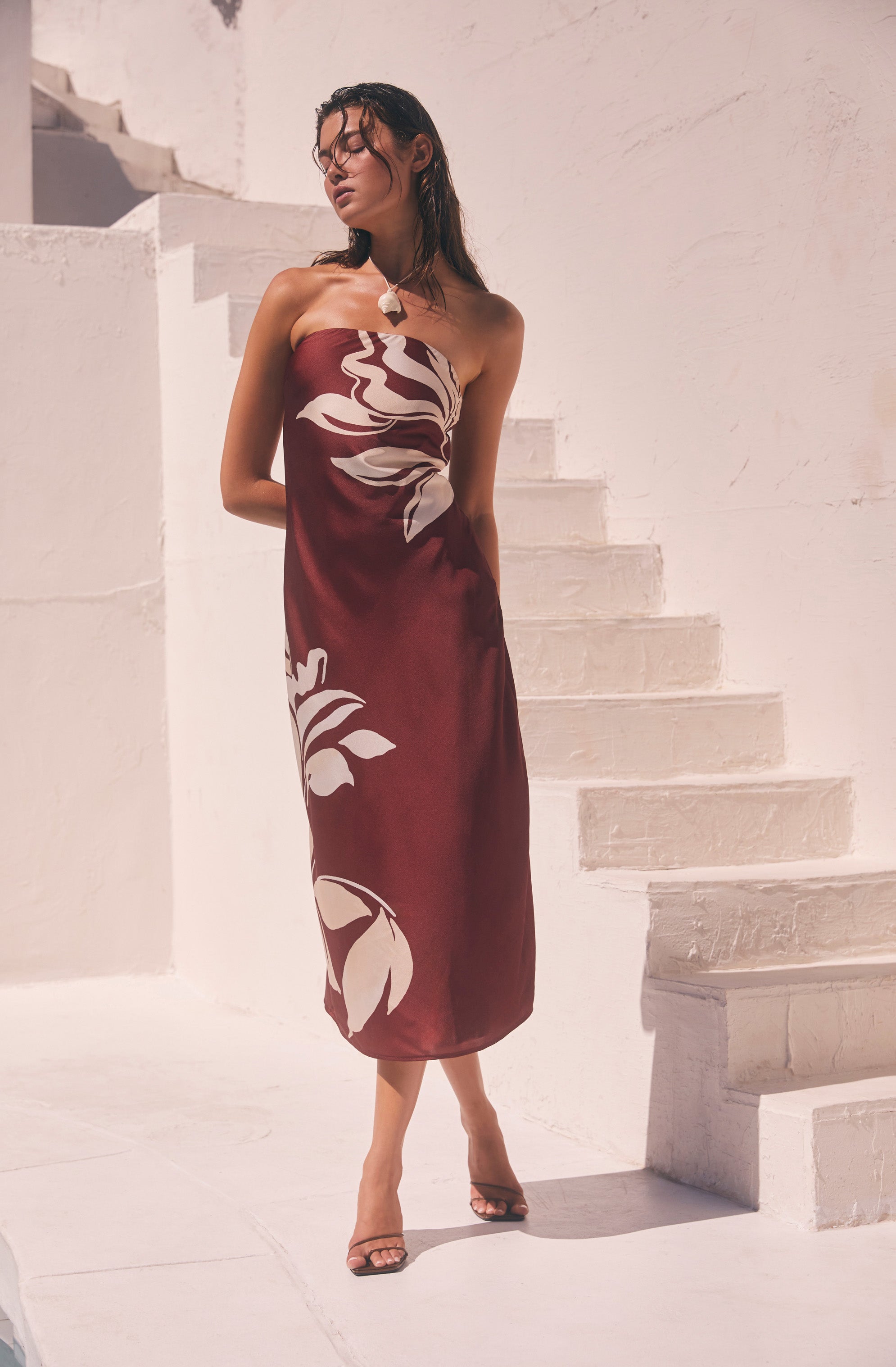 Woman in a strapless, maroon dress with large, cream-colored floral print, standing outdoors near white stairs.