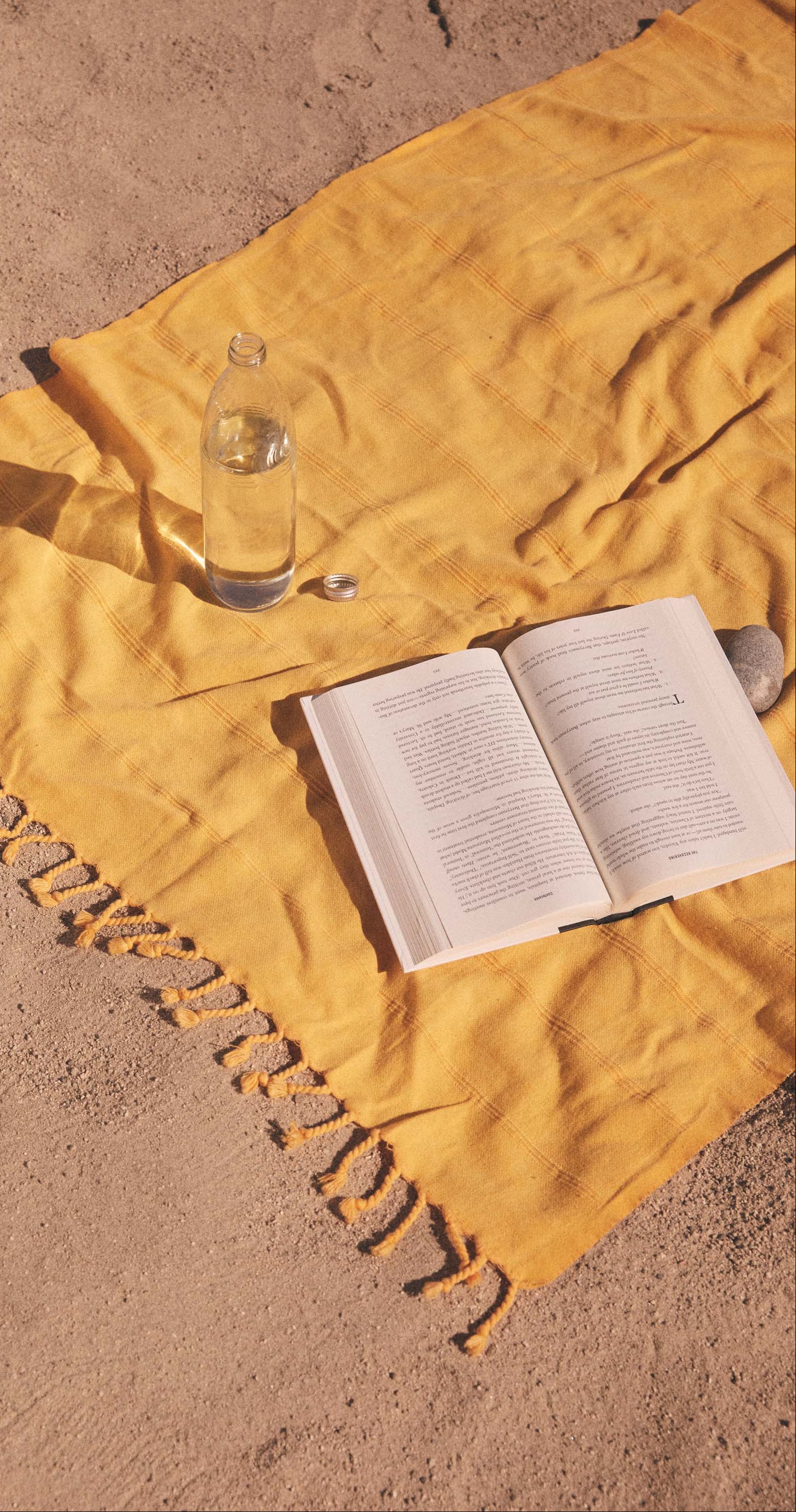 A yellow striped blanket on sand with an open book, a water bottle, and a stone.