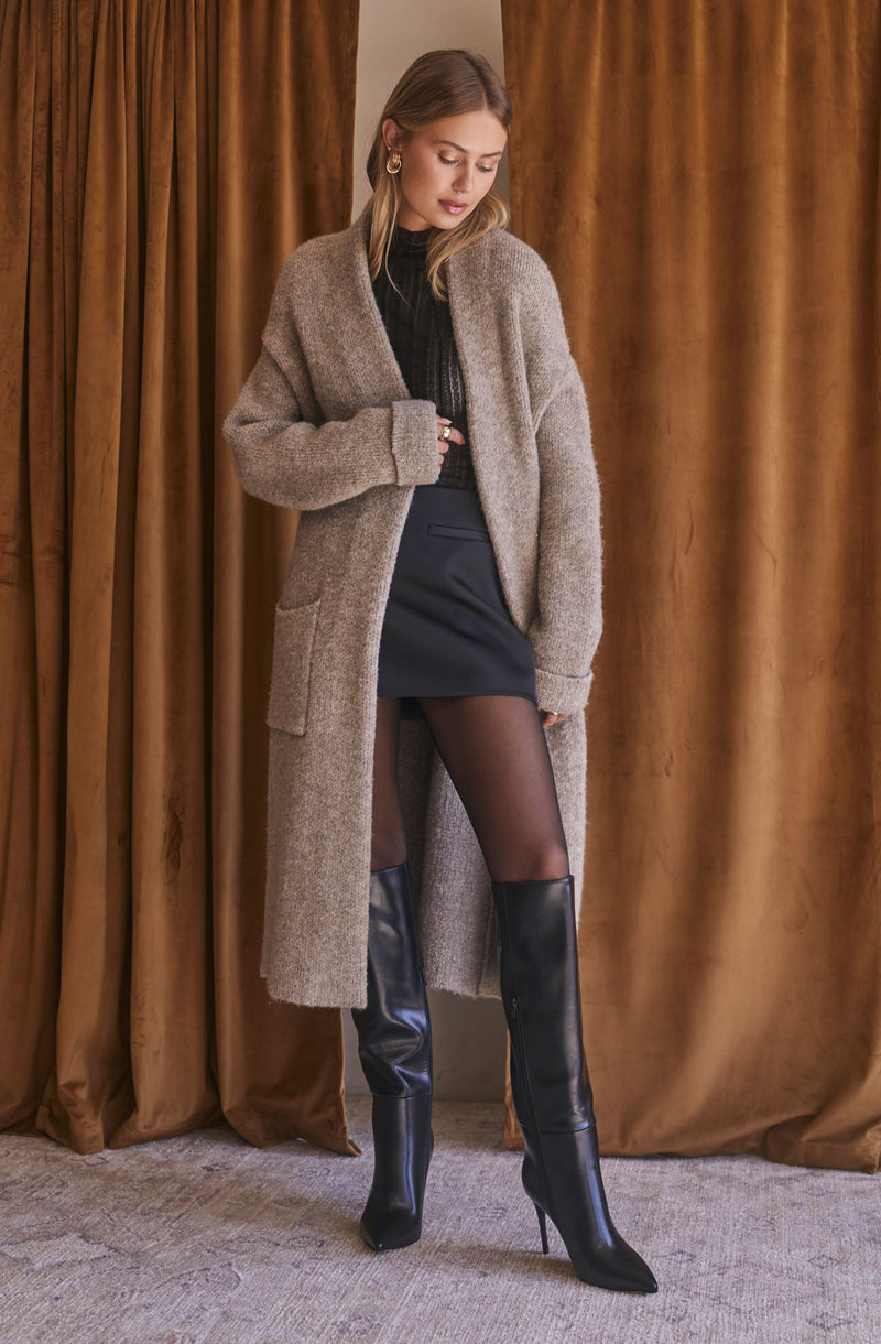 A woman is wearing a long, chunky beige cardigan with large pockets over a black top. She pairs this with a short black skirt, sheer black tights, and tall black knee-high boots with high heels. She is accessorized with large gold hoop earrings. The background features brown velvet curtains.