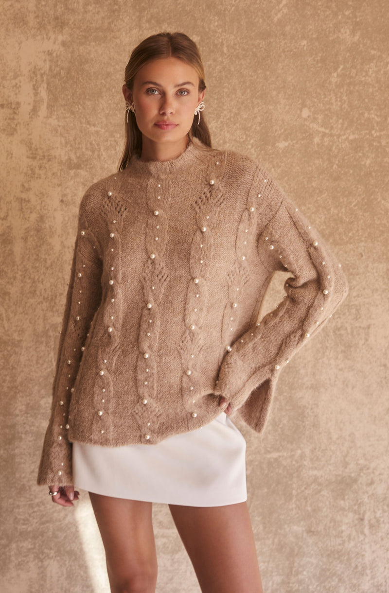 A woman is wearing a loose-knit, beige sweater with a high neckline, decorated with evenly spaced white pearl embellishments. She pairs the sweater with a white skirt. The background is a neutral, textured beige surface.