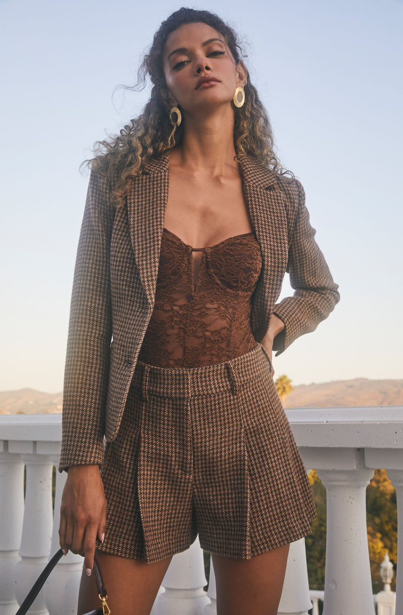 A woman is wearing a brown plaid matching blazer and shorts set. Underneath, she has on a sheer, lace, strapless bodysuit in a matching brown color. She accessorizes with large oval-shaped earrings.