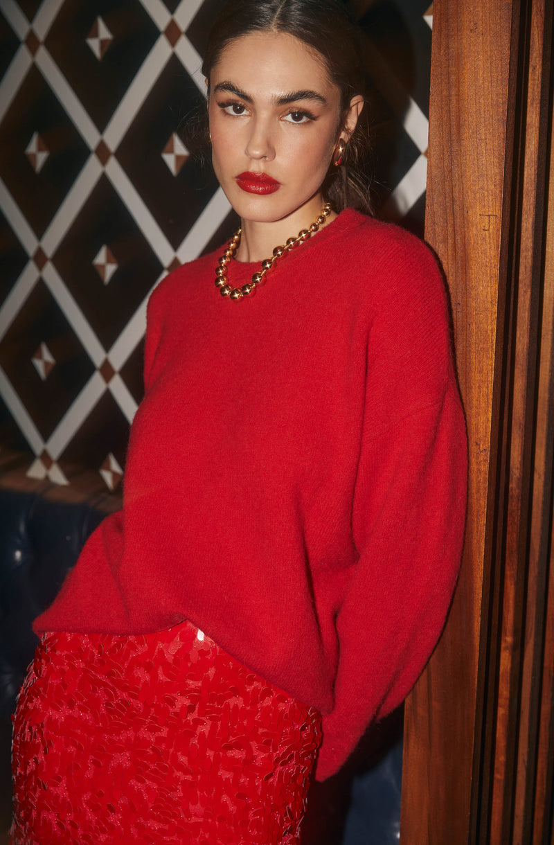 The woman is wearing a bright red, oversized knit sweater with a round neckline. She has paired it with a matching red skirt featuring a textured, abstract pattern. She accessorized with a gold chain necklace and small gold hoop earrings.
