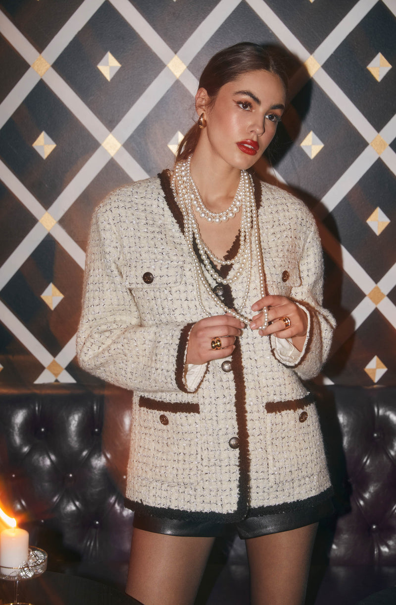 A woman is wearing a cream-colored, textured tweed jacket with dark trim along the edges, pockets, and cuffs. The jacket has a tailored fit with gold buttons. She is accessorized with multiple strands of pearl necklaces, gold earrings, and rings. She is also wearing black shorts made of leather or a similar material. The background features a geometric patterned wall with diamonds and lines in black, white, and gold.