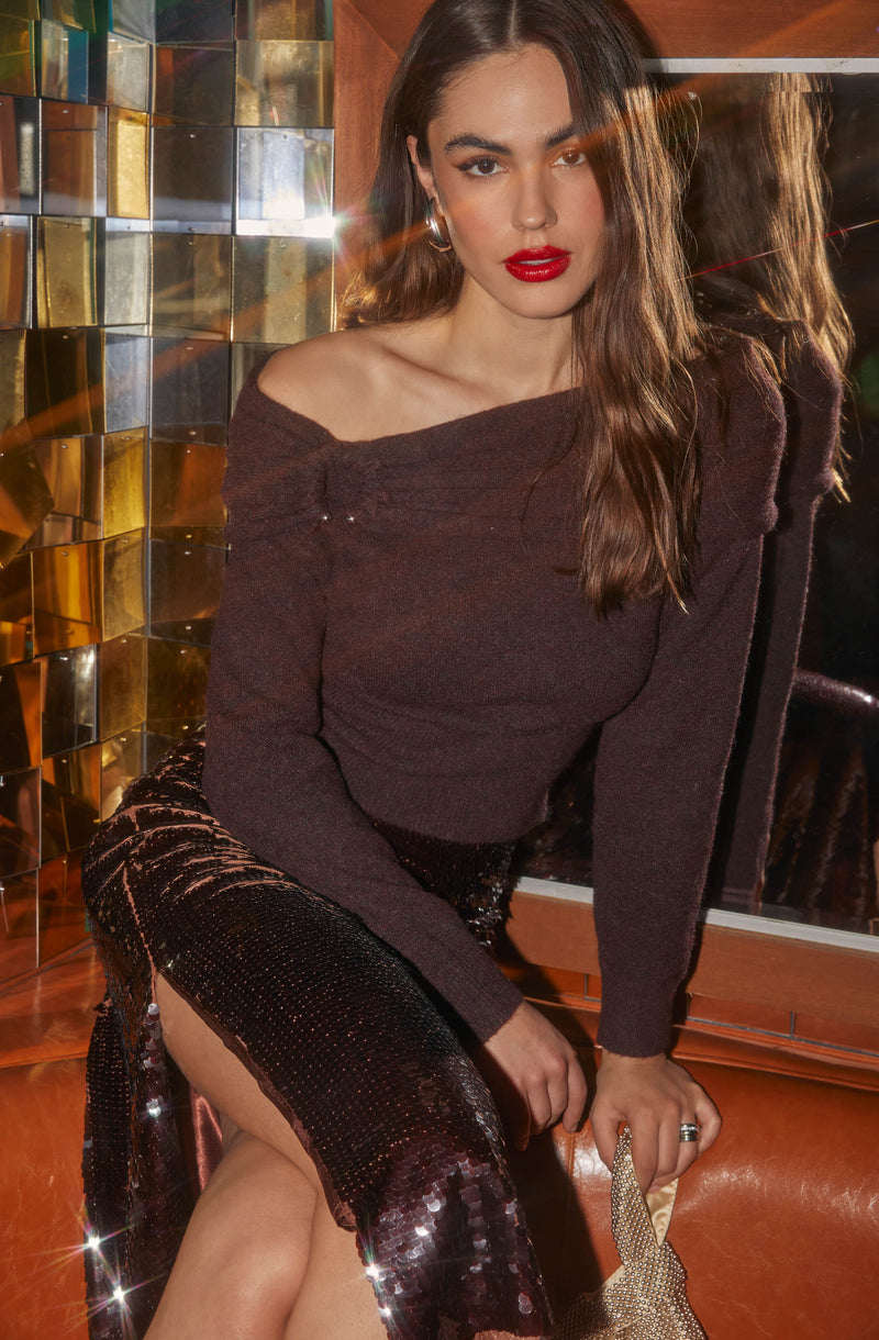 A young woman with long, wavy brown hair is wearing a dark maroon off-the-shoulder sweater that drapes over one shoulder. She has bold red lipstick and hoop earrings. Her skirt is a high-slit, sequined design in dark colors, creating a shimmering, glittering effect. She is holding a small, sparkly clutch bag in her hand. The background features a wall with reflective, multicolored tiles and a mirror that reflects her profile.