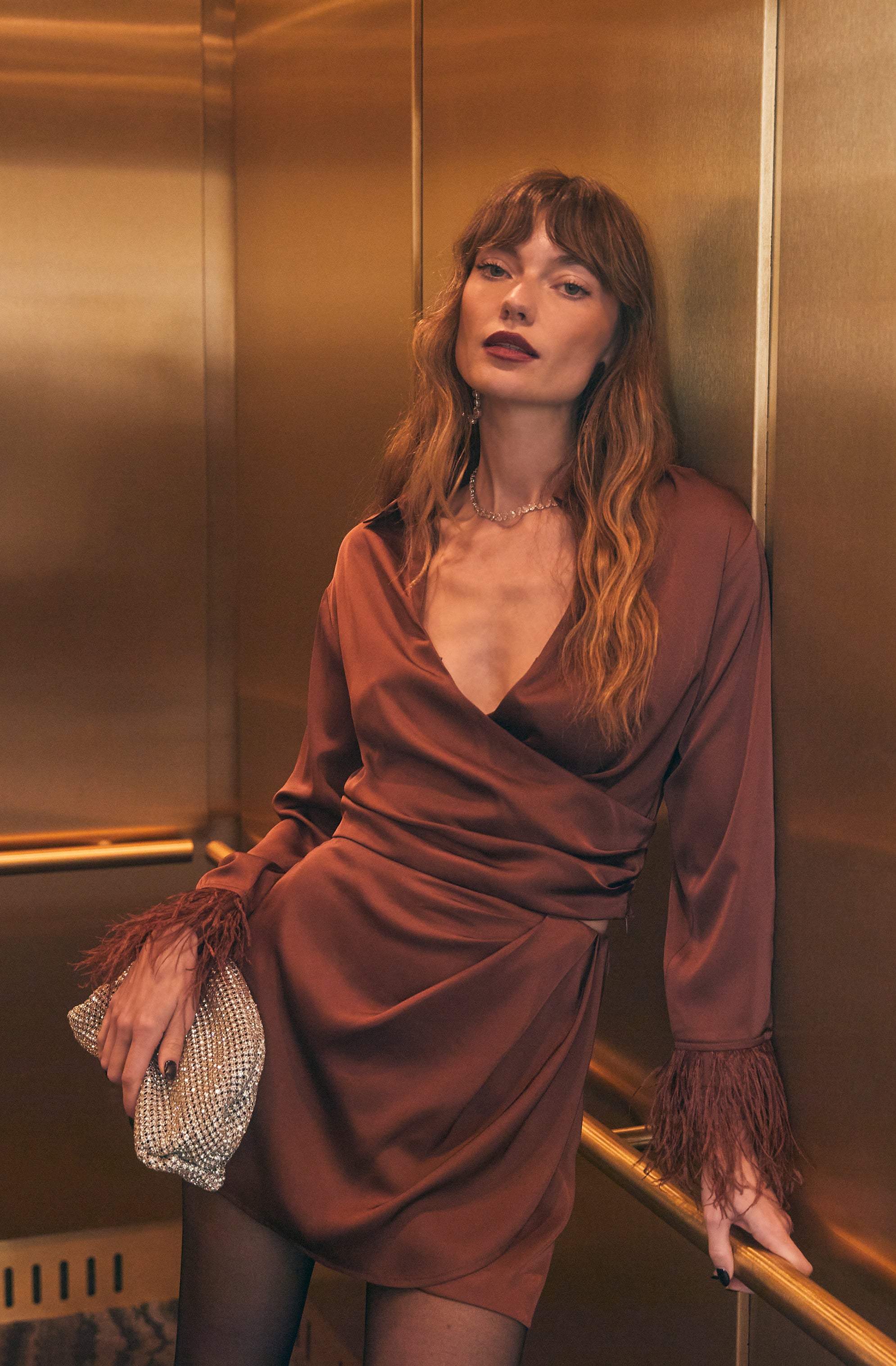 A woman is wearing a satin dress in a rich, deep brown color. The dress has a V-neckline and long sleeves with feathered cuffs. She is holding a shimmering, textured clutch bag and is accessorized with a delicate necklace. Her long, wavy hair is parted in the middle, and she has dark lipstick.