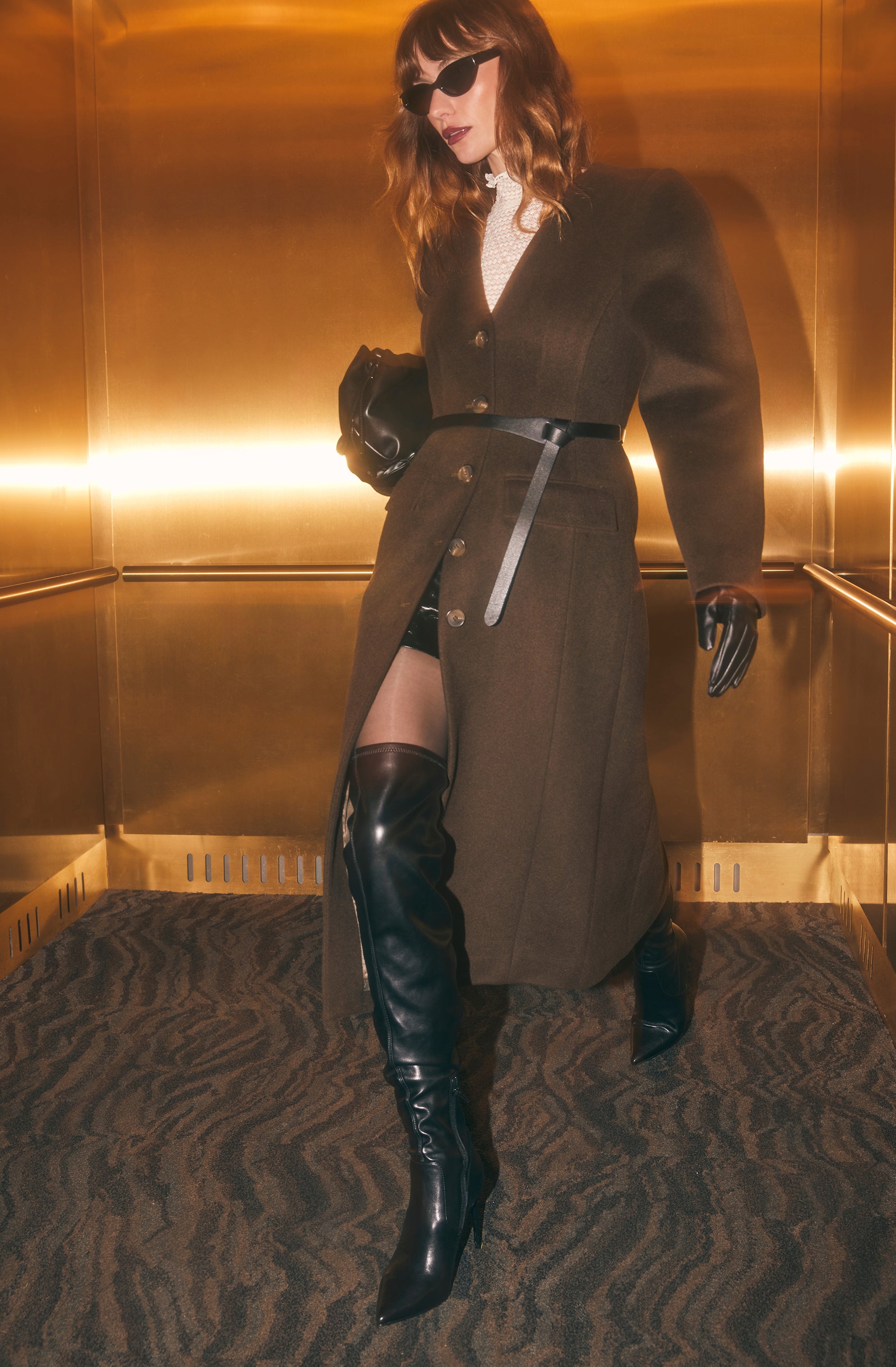 A woman is wearing a long, dark brown coat that is buttoned in the front and cinched at the waist with a black belt. Underneath, she has on a white top with a textured pattern. She is also wearing black, thigh-high leather boots with pointed toes and stiletto heels, paired with black leather gloves. She has on dark sunglasses and has wavy brown hair. The background features a metallic, gold-colored wall and lighting.