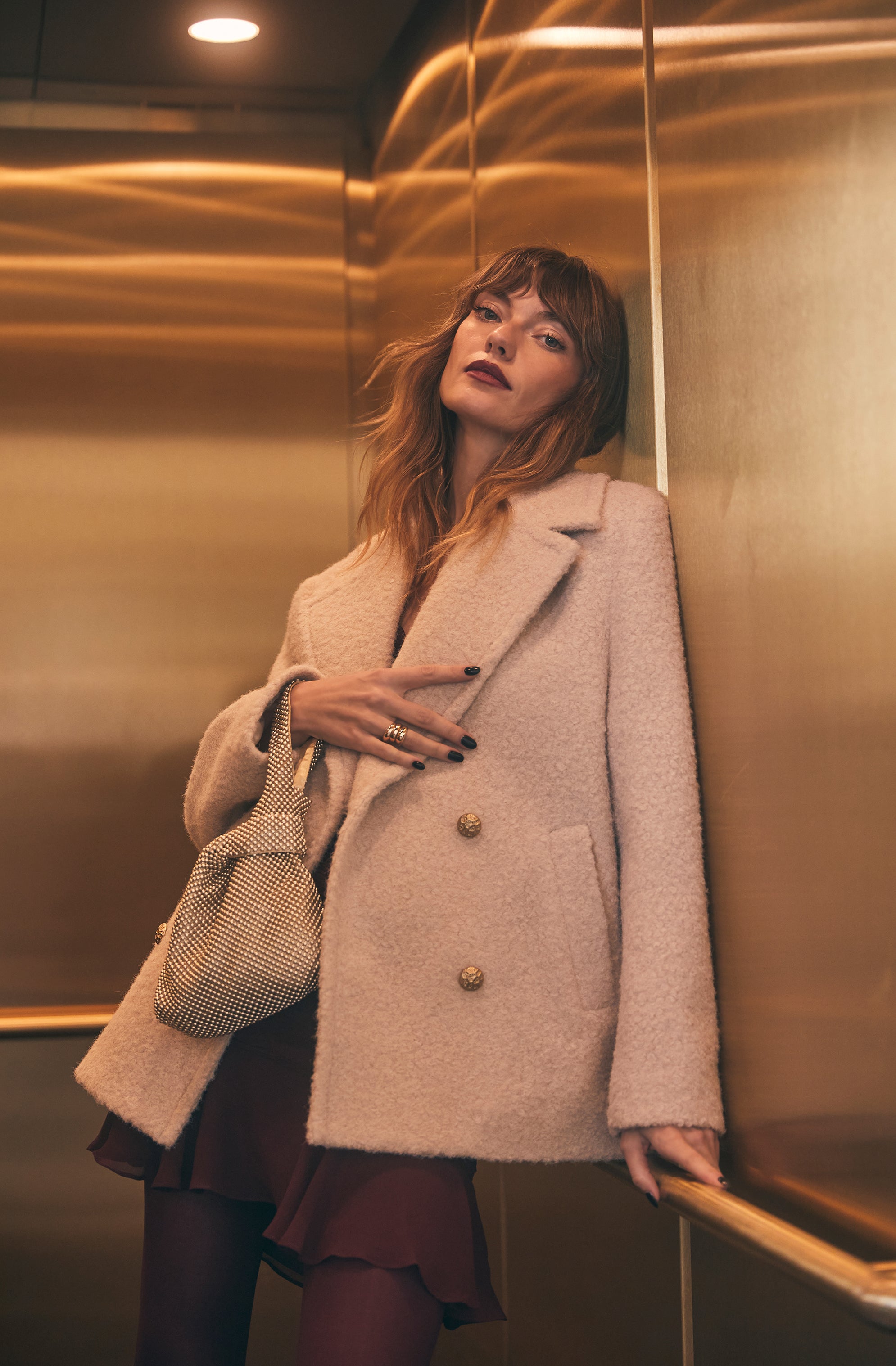 A woman is wearing a pale pink, double-breasted, oversized coat with gold buttons, paired with dark tights and a dark dress or skirt underneath. She is also carrying a small, beige, patterned handbag over her shoulder.