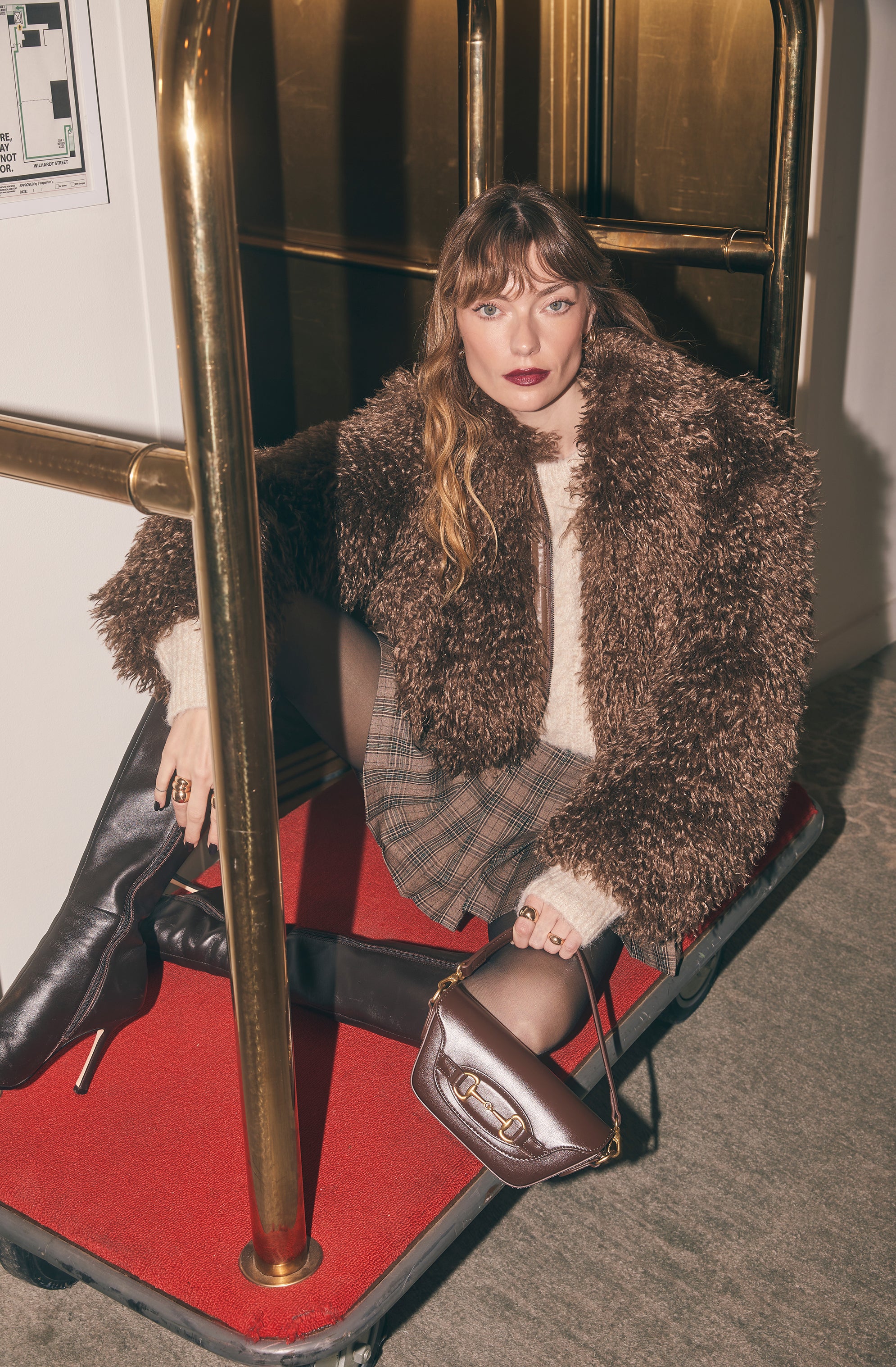 A woman is seated on a luggage cart inside a building. She is wearing a plush, brown, fluffy coat over a white sweater. Her outfit includes a plaid skirt with shades of brown, beige, and red, paired with black sheer tights. She is also wearing black knee-high heeled boots. She is holding a small brown handbag with a gold chain strap, and there are multiple rings on her fingers. Her makeup features dark lipstick, and she has long wavy hair with bangs.