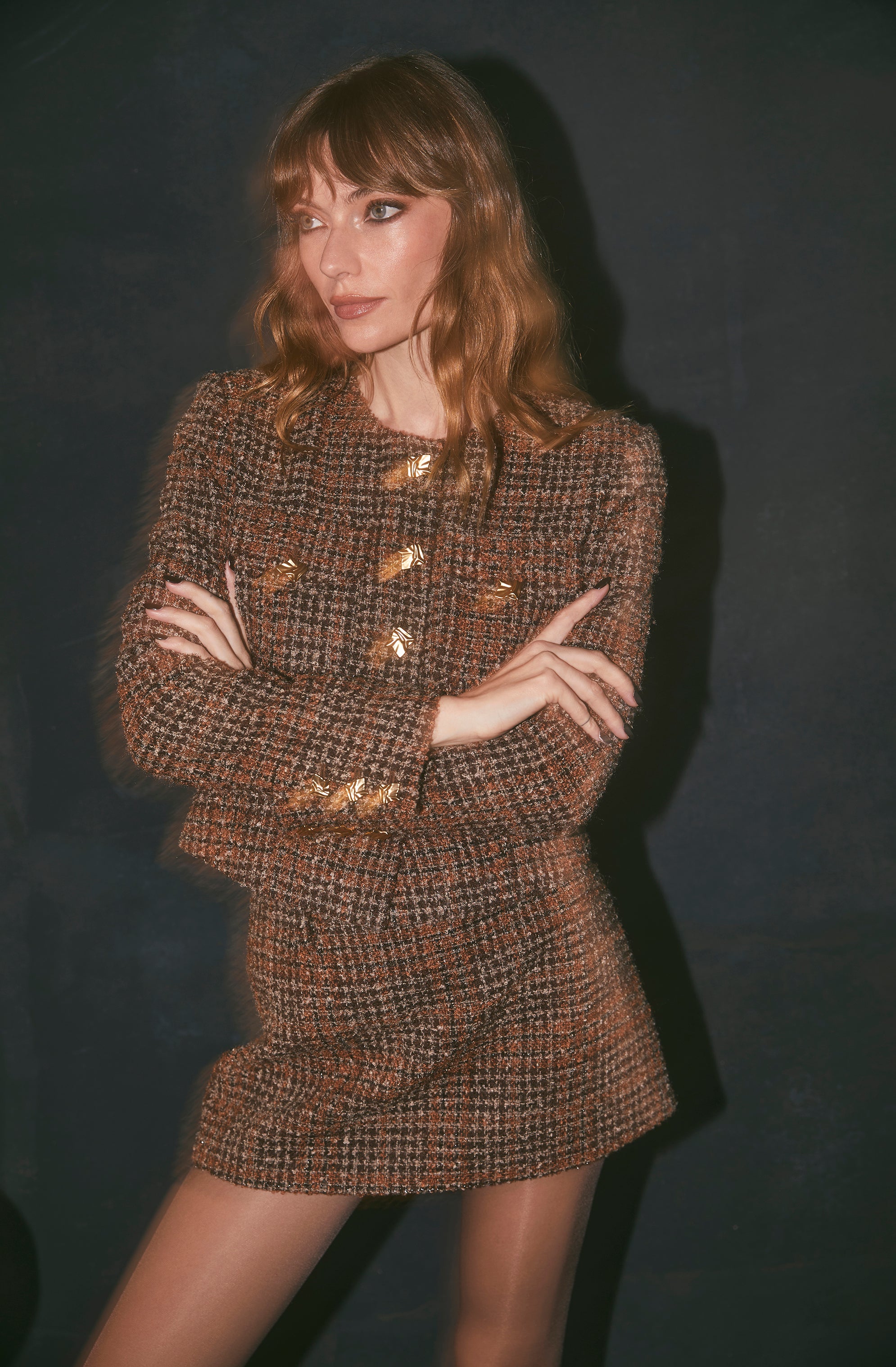A woman is wearing a matching two-piece outfit made of brown, black, and orange houndstooth tweed fabric. The top is a fitted jacket with a round neckline, decorated with large gold buttons down the front and on the cuffs. The skirt is a high-waisted, short, fitted mini skirt made from the same fabric. She has fair skin, light makeup, and shoulder-length wavy brown hair with blunt bangs.