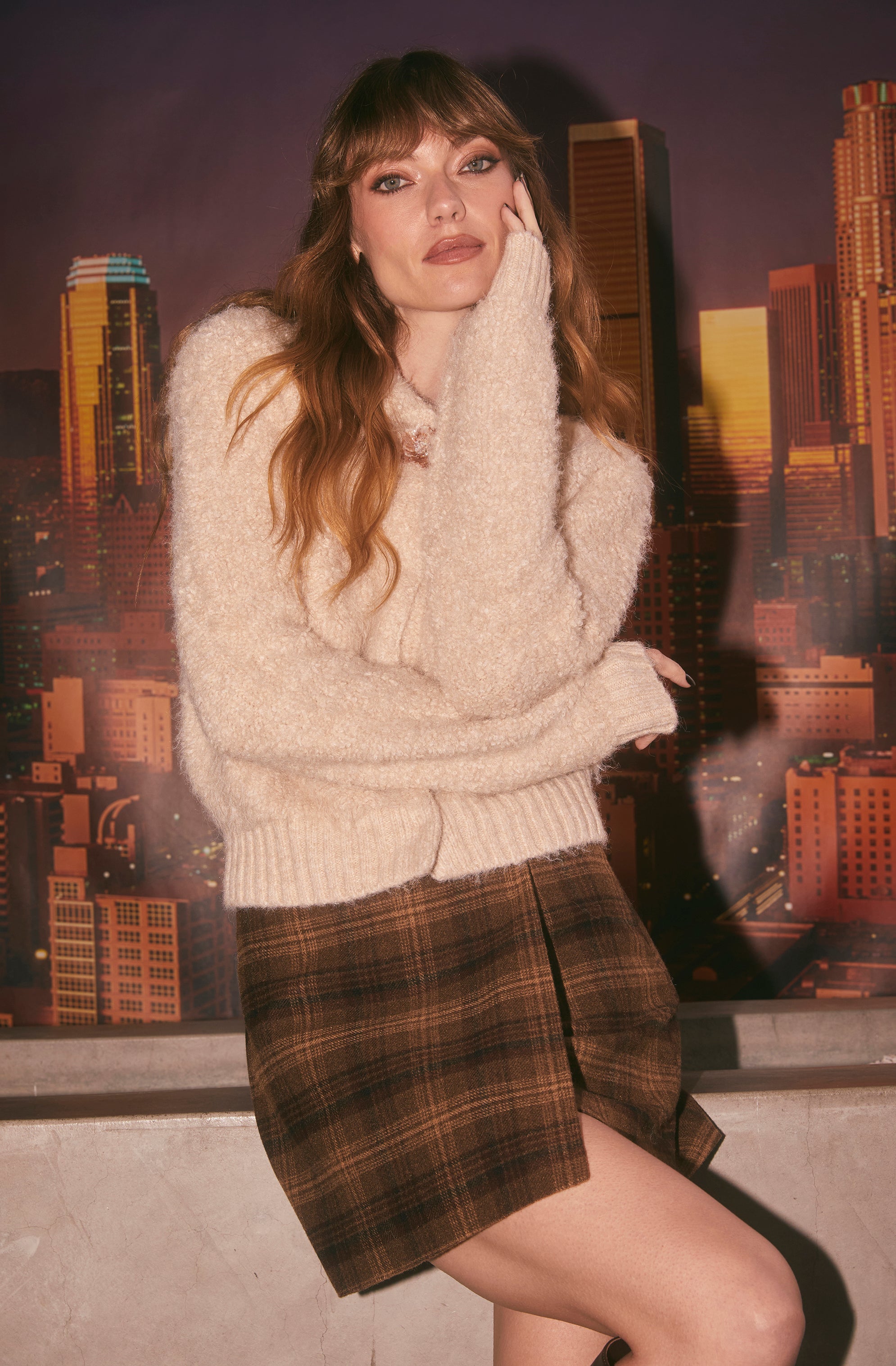 The woman is wearing a fluffy, cream-colored sweater with long sleeves and a ribbed hem. She is also dressed in a high-waisted, knee-length plaid skirt in shades of brown and beige.