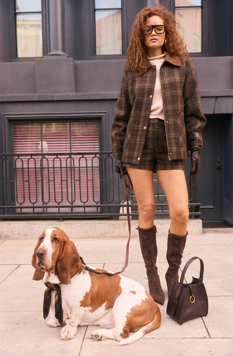 Woman in plaid jacket and shorts, wearing knee-high boots and gloves, standing outdoors with a Basset Hound.