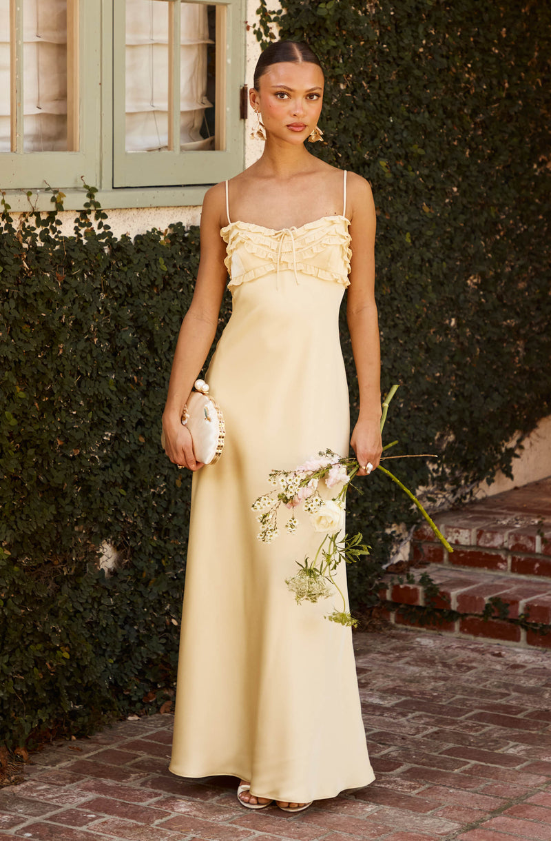 Woman in a pale yellow, ruffled, spaghetti strap maxi dress holding a bouquet of flowers.