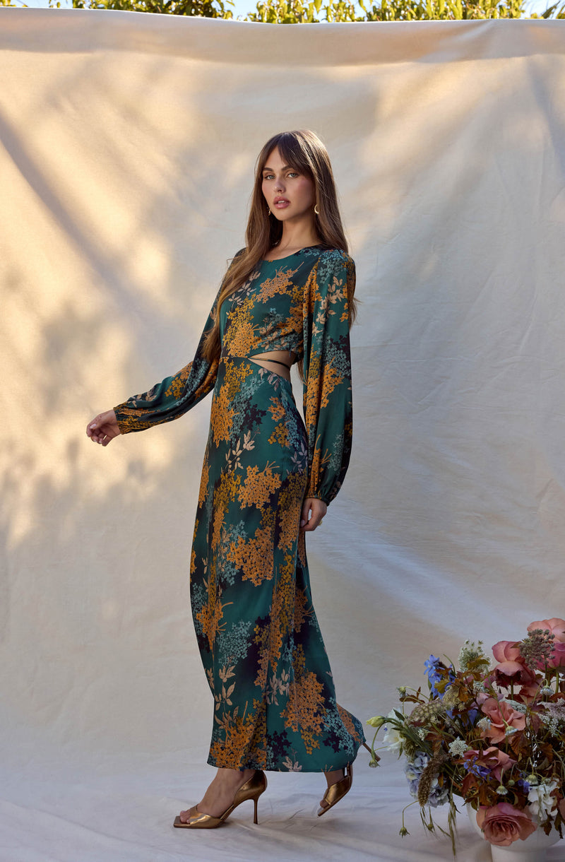 A woman is wearing a long, flowing dress with a dark green base color and a floral pattern in shades of yellow, orange, and beige. The dress has long, billowy sleeves and features a cut-out detail at the waist. She is also wearing a pair of open-toe, high-heeled shoes in a metallic gold color.