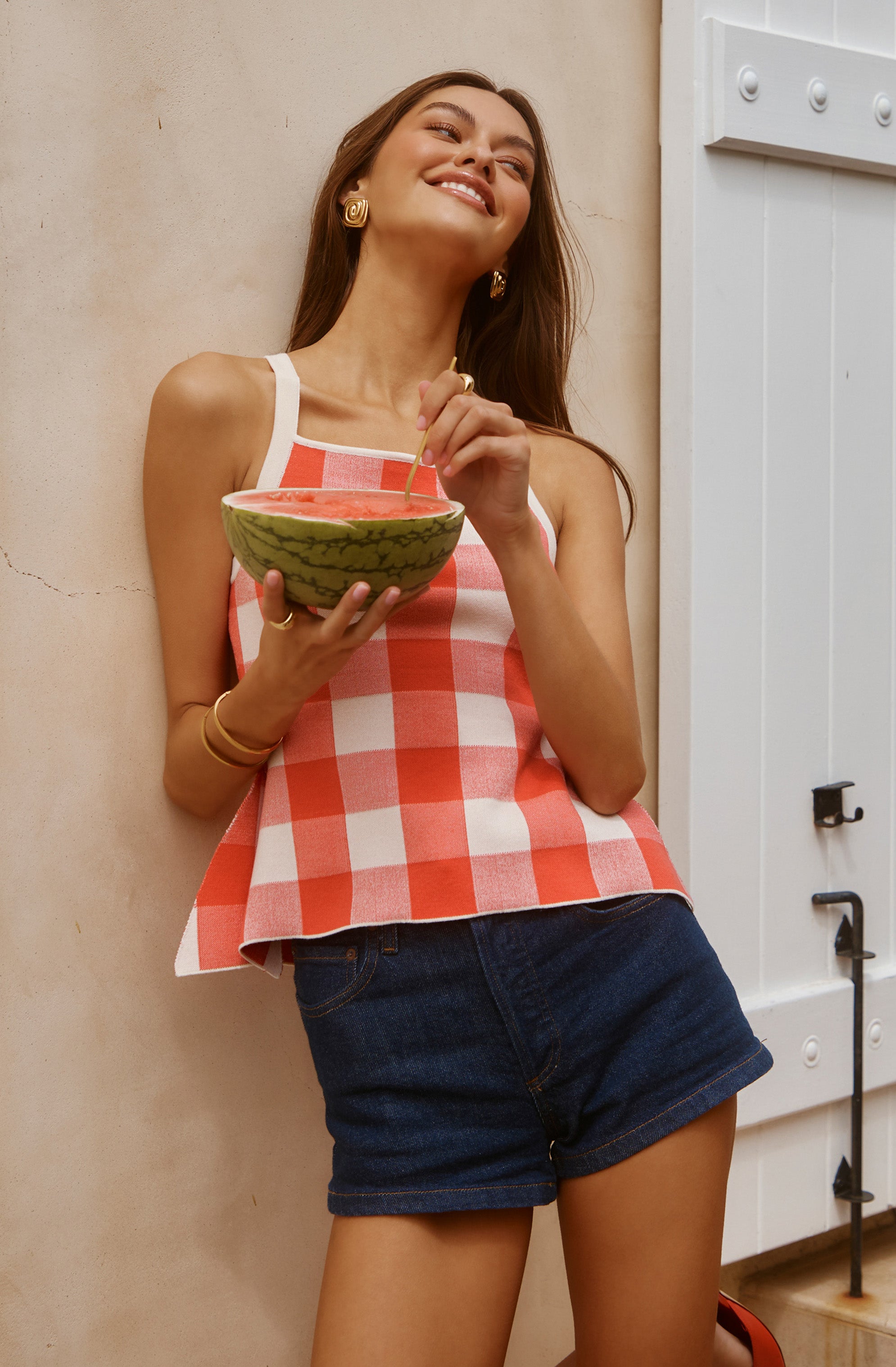Woman in a red and white checkered sleeveless top holding a watermelon slice, wearing dark denim shorts and gold jewelry.