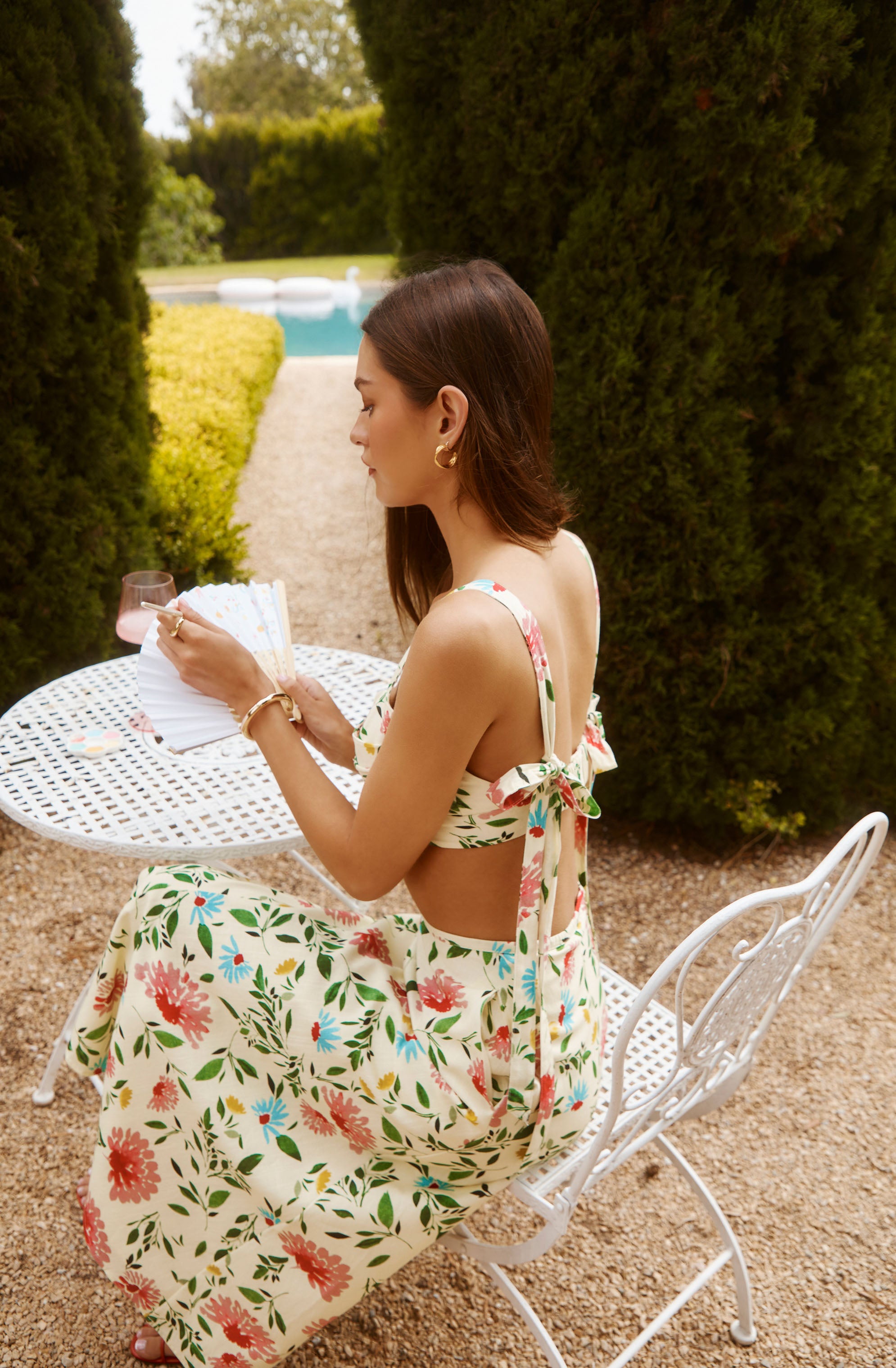 Woman in floral cropped top with bow tie straps and matching flowy skirt, sitting outdoors by a white metal table.