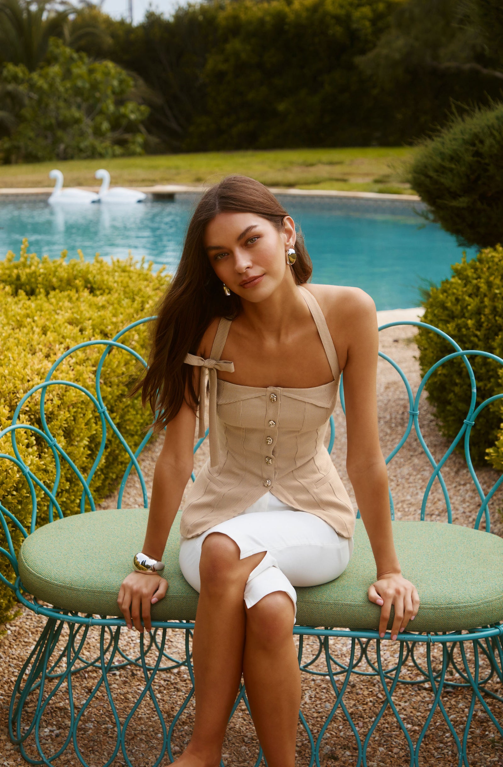 Woman in beige sleeveless top and white pants sitting on a green cushioned bench outdoors near a pool.
