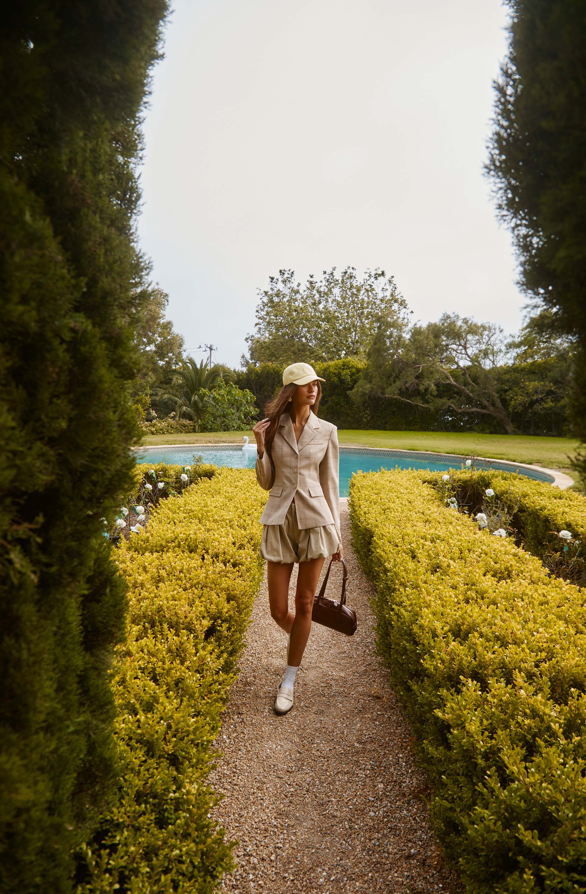 Woman in beige blazer, shorts, white sneakers, and cream cap walking along a garden path.