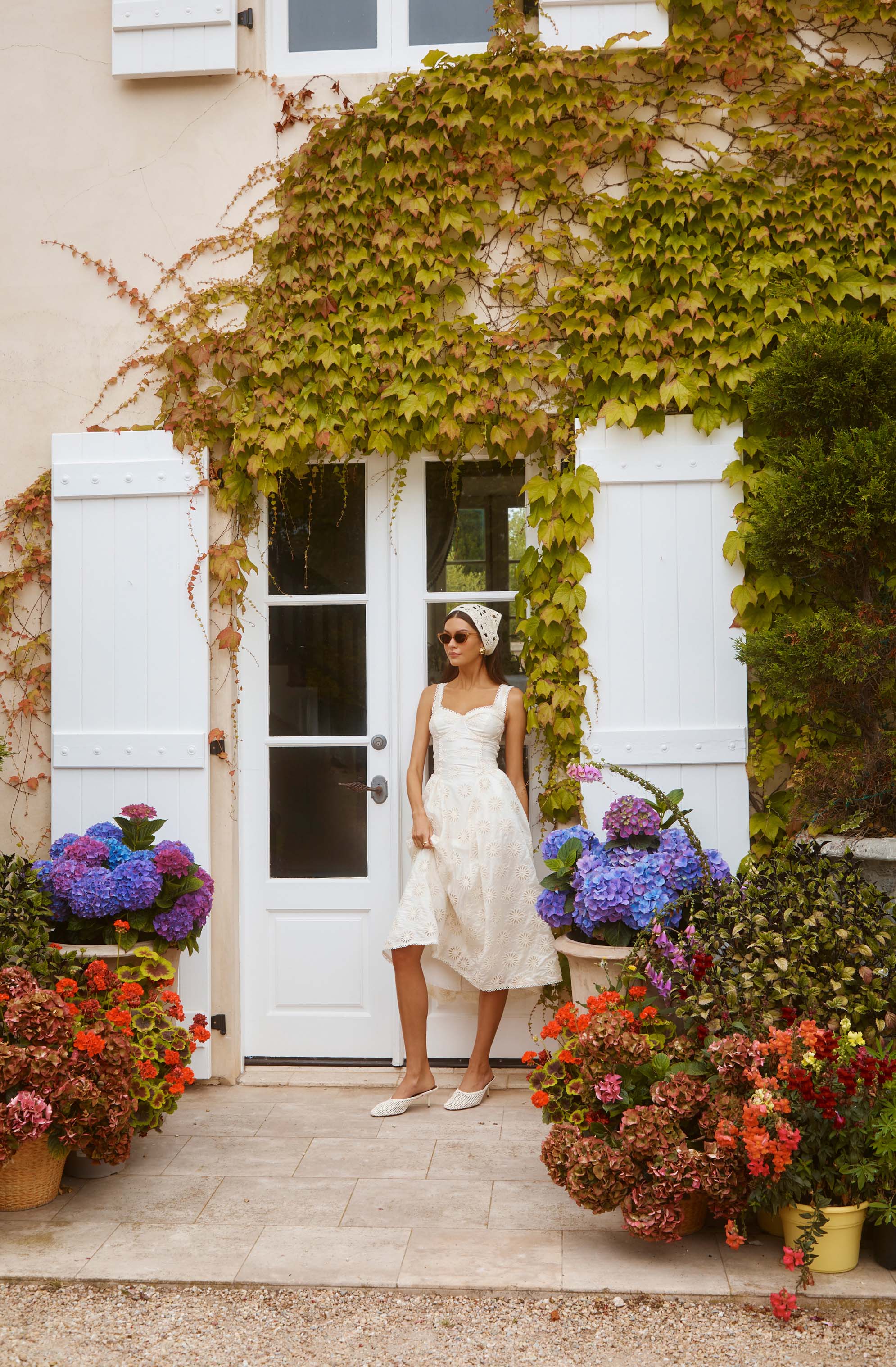 Woman in white dress and leopard-print headscarf, standing by a white door with green vines and colorful flowers outside.