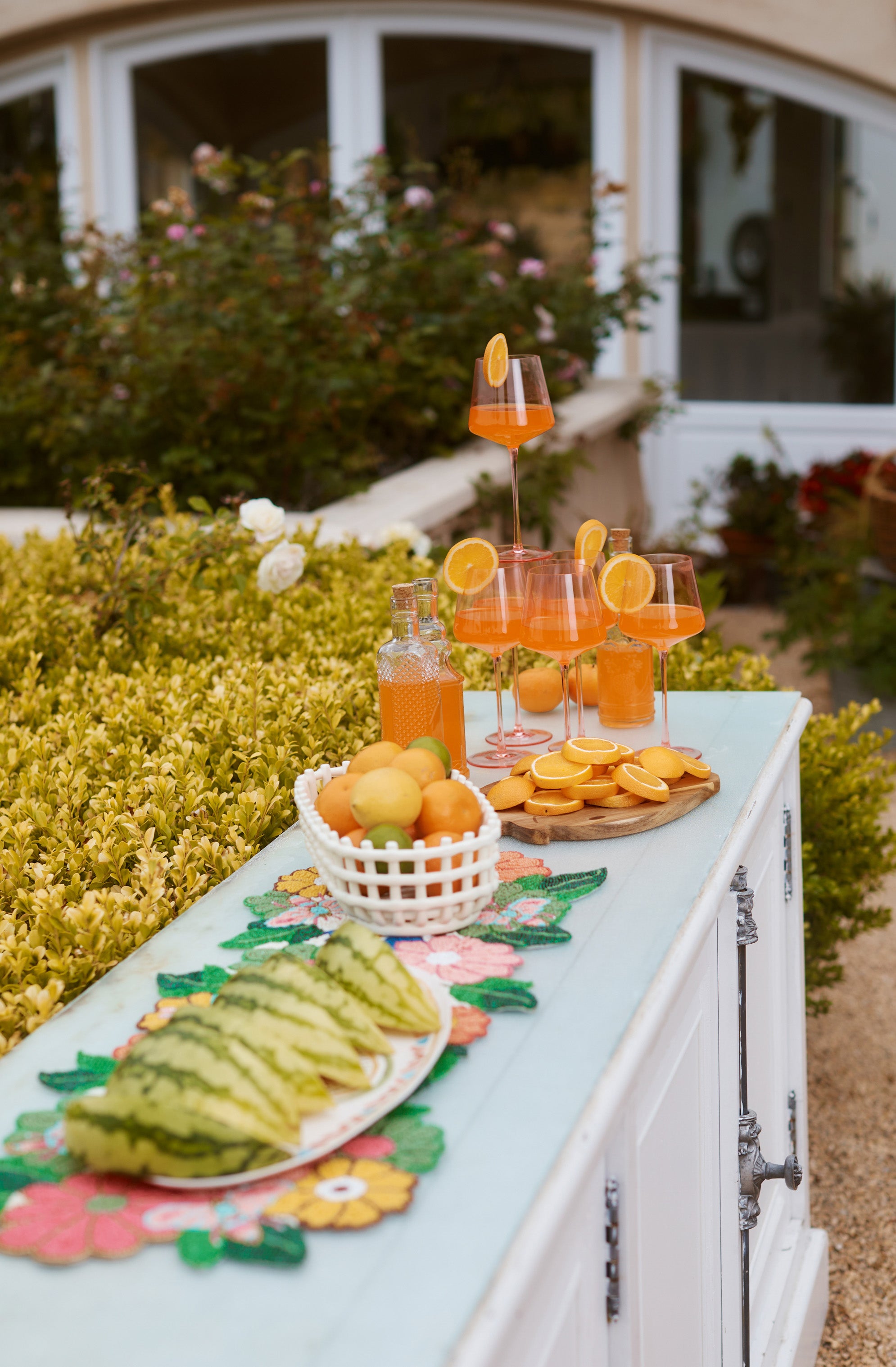 No people; focus on the outdoor table with drinks, lemon slices, and sliced watermelon on a colorful floral cloth.