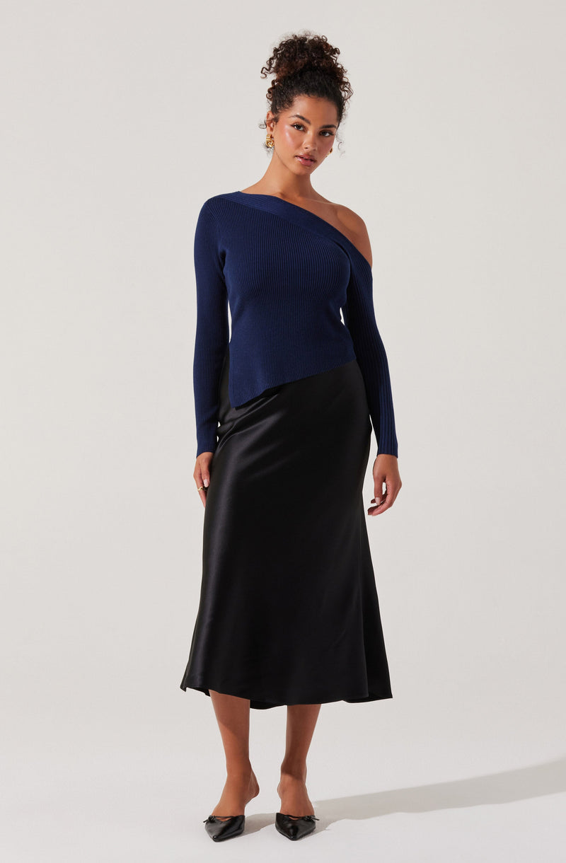 A woman is wearing a dark navy blue, asymmetrical, off-the-shoulder knitted sweater with a ribbed texture and a longer sleeve on one side. She pairs it with a black, midi-length, shiny satin slip skirt. She is also wearing black pointed-toe heels and gold earrings.