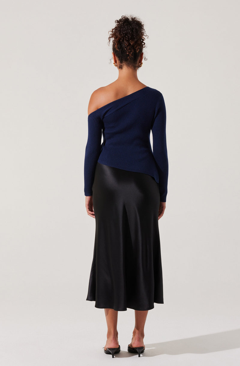A woman is wearing a dark blue, off-the-shoulder knit sweater with long sleeves. The sweater has a ribbed texture and is asymmetrical, draping off one shoulder. She is also wearing a black satin midi skirt with a slight sheen and a flowing silhouette. She has curly hair styled up and is standing against a plain light background.