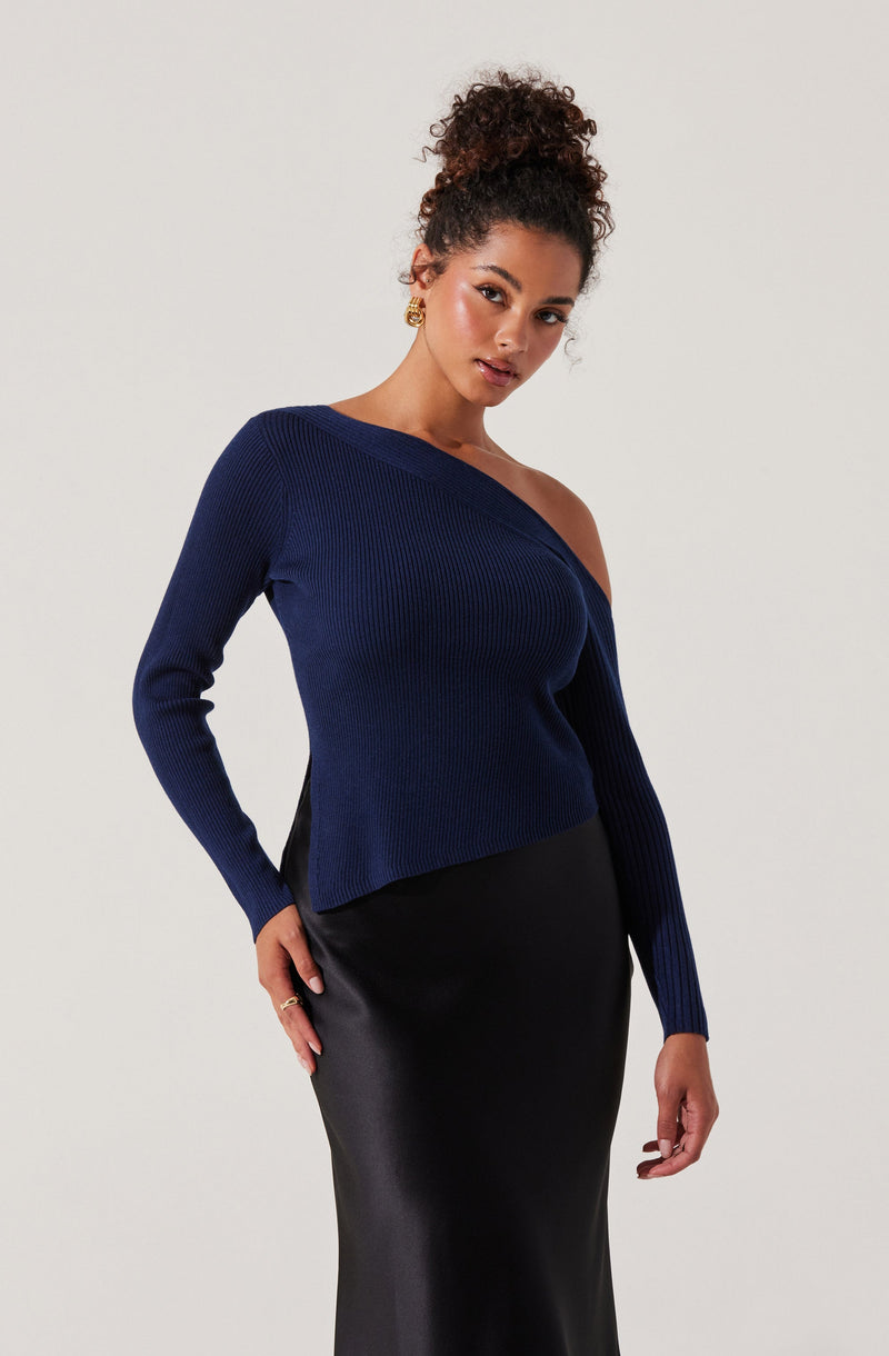 A woman is wearing a dark blue, ribbed, off-the-shoulder sweater with long sleeves. She pairs it with a black, fitted, leather-like skirt. She has styled her hair in a full, curly bun and is accessorized with gold earrings and rings.
