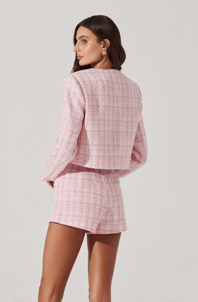 A woman is wearing a light pink, textured, checkered two-piece set consisting of a cropped blazer with long sleeves and matching high-waisted shorts. The blazer has a boxy fit with no visible collar.