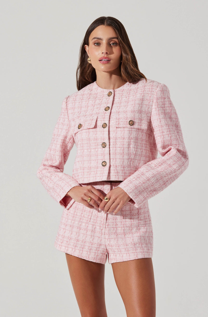 A woman is wearing a light pink, textured, tweed-like suit consisting of a cropped blazer and matching high-waisted shorts. The blazer features a round neckline, long sleeves, and decorative gold buttons down the front. It also has two small flap pockets on the chest with gold buttons.