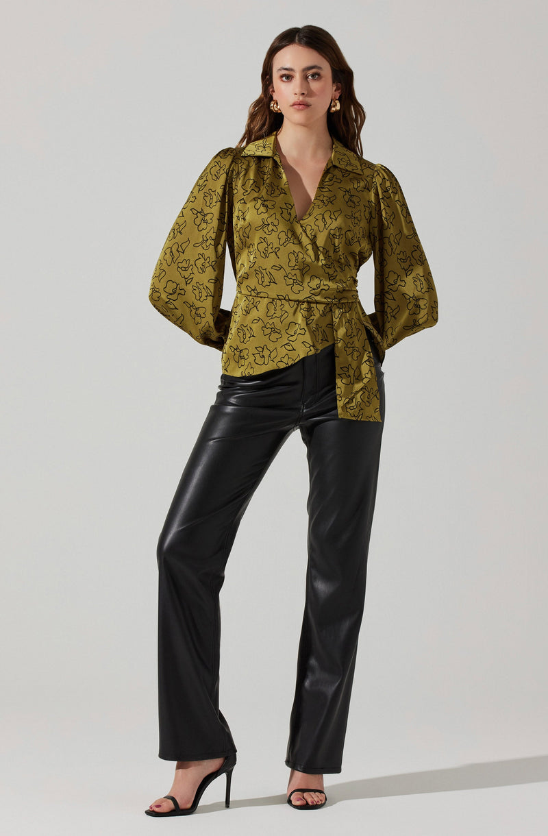 A woman is wearing a silky, olive-green blouse with a black abstract floral pattern, featuring a collar, a deep V-neckline, and puffed long sleeves that are gathered at the cuffs. The blouse is cinched at the waist with a matching fabric belt tied in a knot. She pairs it with high-waisted, black, faux leather wide-leg pants and black high-heeled sandals with thin straps. She has gold hoop earrings and her hair is styled in loose waves.