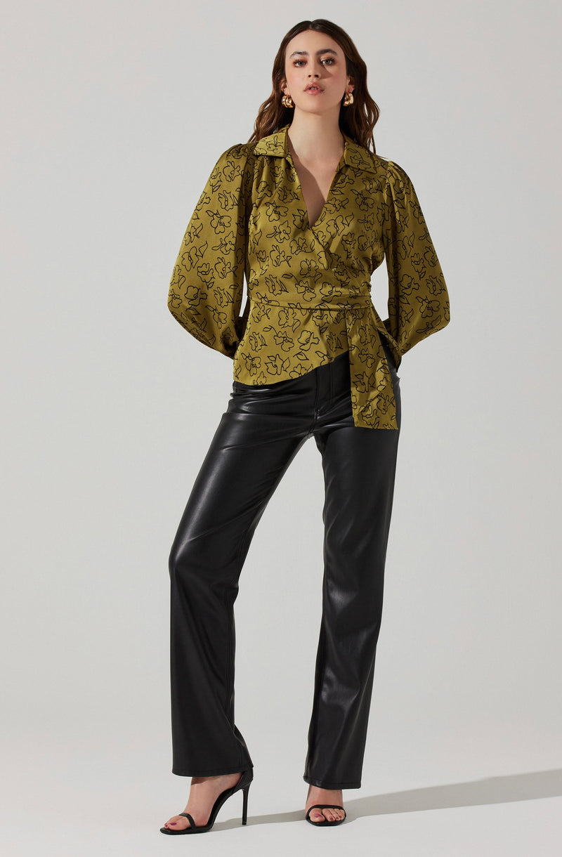 A woman is wearing a long-sleeved, olive-green blouse with black floral outlines, featuring a collar and a wrap-around tie at the waist. She pairs the blouse with black faux leather pants and black high-heeled sandals with ankle straps. She is also accessorized with gold hoop earrings.