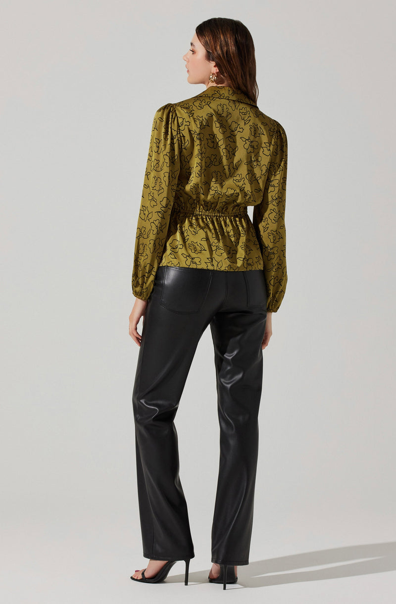 A woman is wearing a long-sleeved, olive green blouse with a black floral outline pattern. The blouse features a gathered waistline and puffed sleeves. She pairs it with high-waisted, black leather pants that have a straight-leg fit. She completes her outfit with black high-heeled sandals.