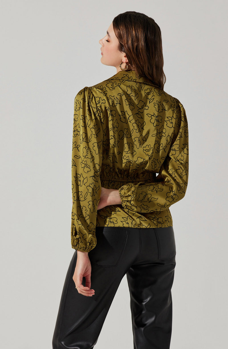A woman is wearing a long-sleeve, olive-green blouse with a black floral outline pattern. The blouse has gathered cuffs and an elastic waistband, creating a slightly bloused effect at the waist. She is also wearing high-waisted black pants made of a shiny, leather-like material.
