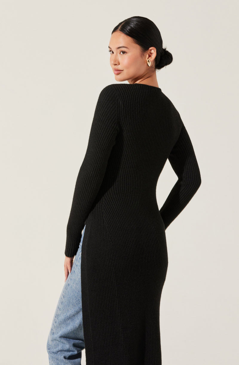 A woman wearing a long black ribbed knit sweater dress with a high neckline. The dress fits snugly and extends down past her knees. She pairs it with light blue high-waisted jeans.