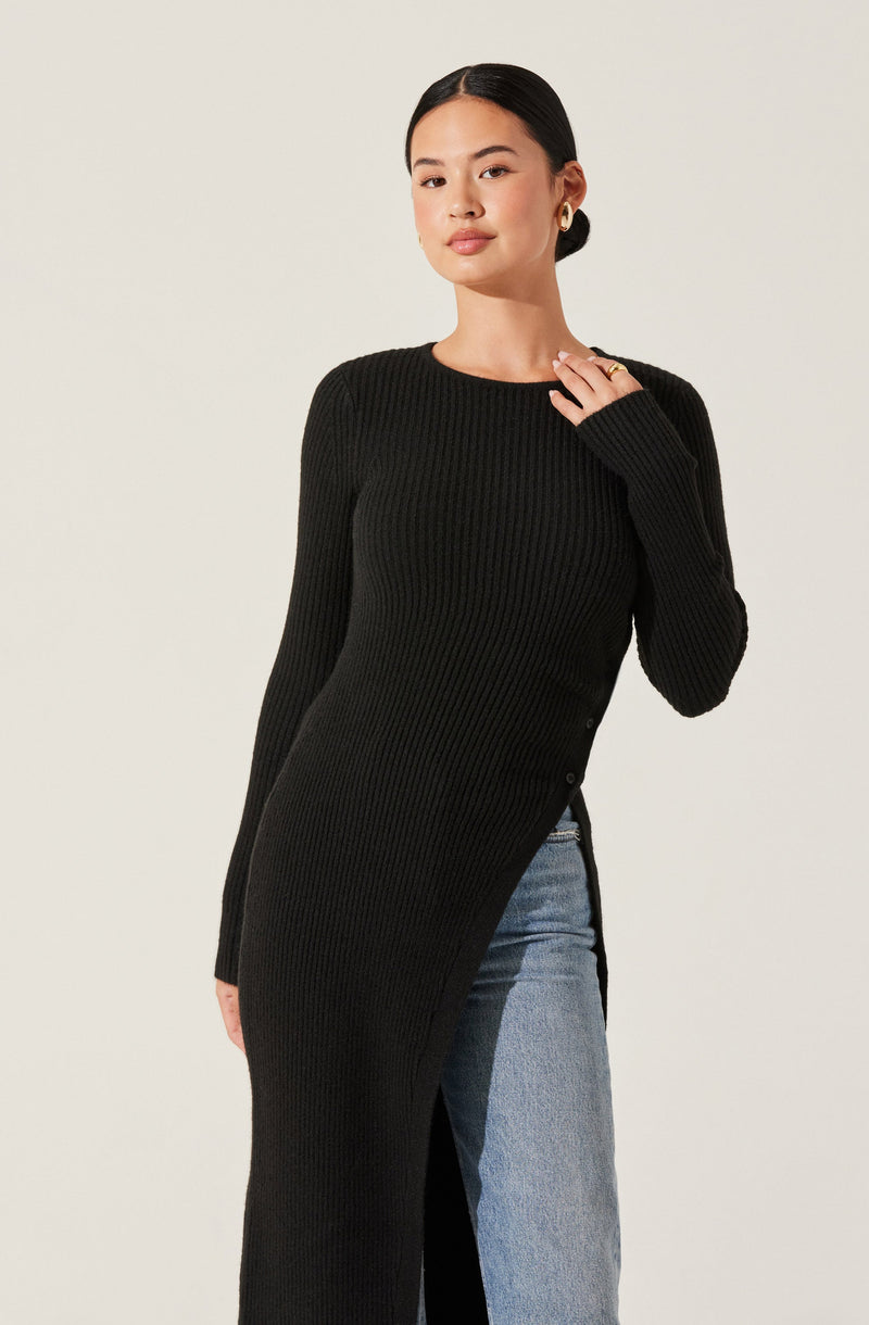 A woman is wearing a black, long-sleeved, ribbed knit dress with a side slit. The dress has a crew neckline and a fitted silhouette. She pairs it with blue jeans visible through the slit. She is accessorized with gold stud earrings and has her hair pulled back.