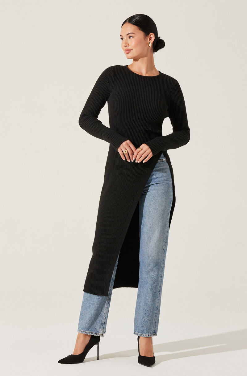 A woman is wearing a long black ribbed knit sweater that extends to her ankles with a side slit. She pairs it with light blue, high-waisted, straight-leg jeans with a slightly faded wash. She completes the outfit with black high-heeled pumps and simple gold hoop earrings.