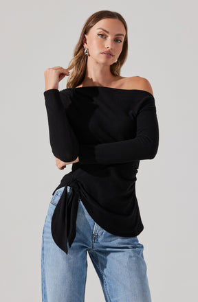 A woman is wearing a black off-the-shoulder long-sleeve top with a side tie detail, paired with light-wash blue jeans.