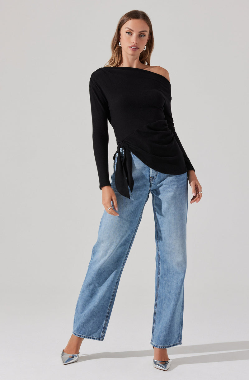 A woman is wearing a black off-the-shoulder long-sleeve top with a side tie detail. She is also wearing light-wash, wide-leg jeans. She has on silver pointed-toe high heels and is accessorized with earrings and rings.