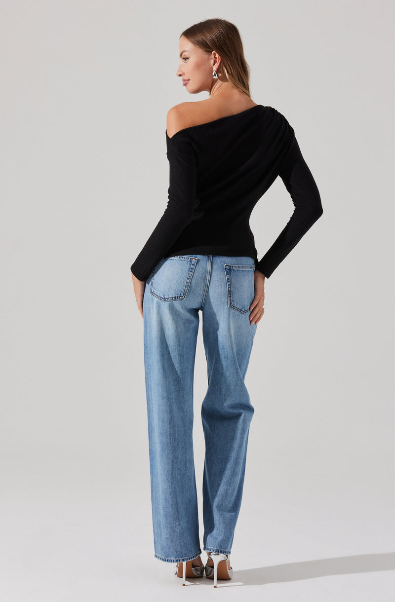 A woman is wearing a black off-the-shoulder sweater with long sleeves and light blue, high-waisted, wide-leg jeans. She has on white high-heeled shoes and is accessorized with statement earrings. She is standing against a plain, light background.