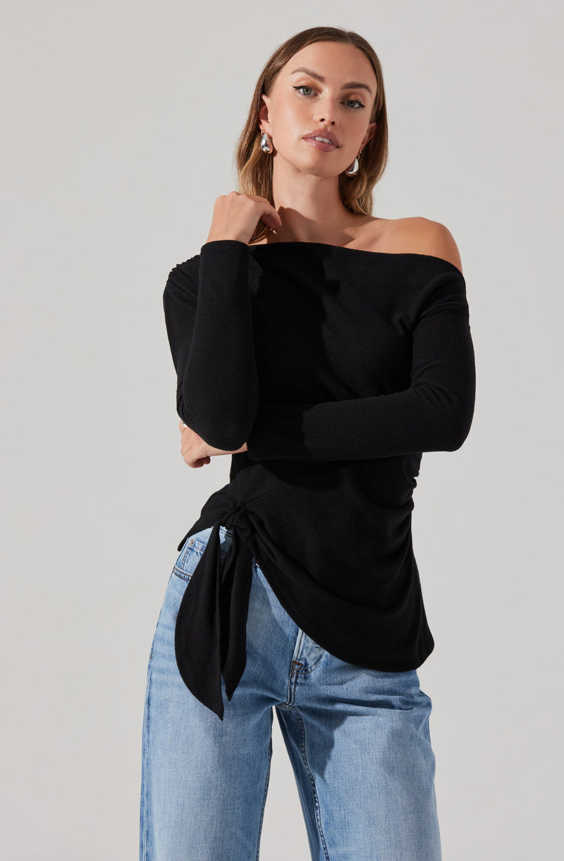 A woman is wearing an off-the-shoulder black top with long sleeves and a waist tie, paired with light-wash blue jeans.