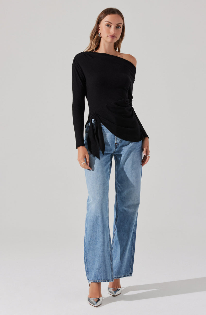 A woman is wearing a black, asymmetrical off-the-shoulder sweater with a knot detail on one side, paired with light blue wide-leg jeans. She completes the look with pointed-toe high heels featuring a metallic accent.
