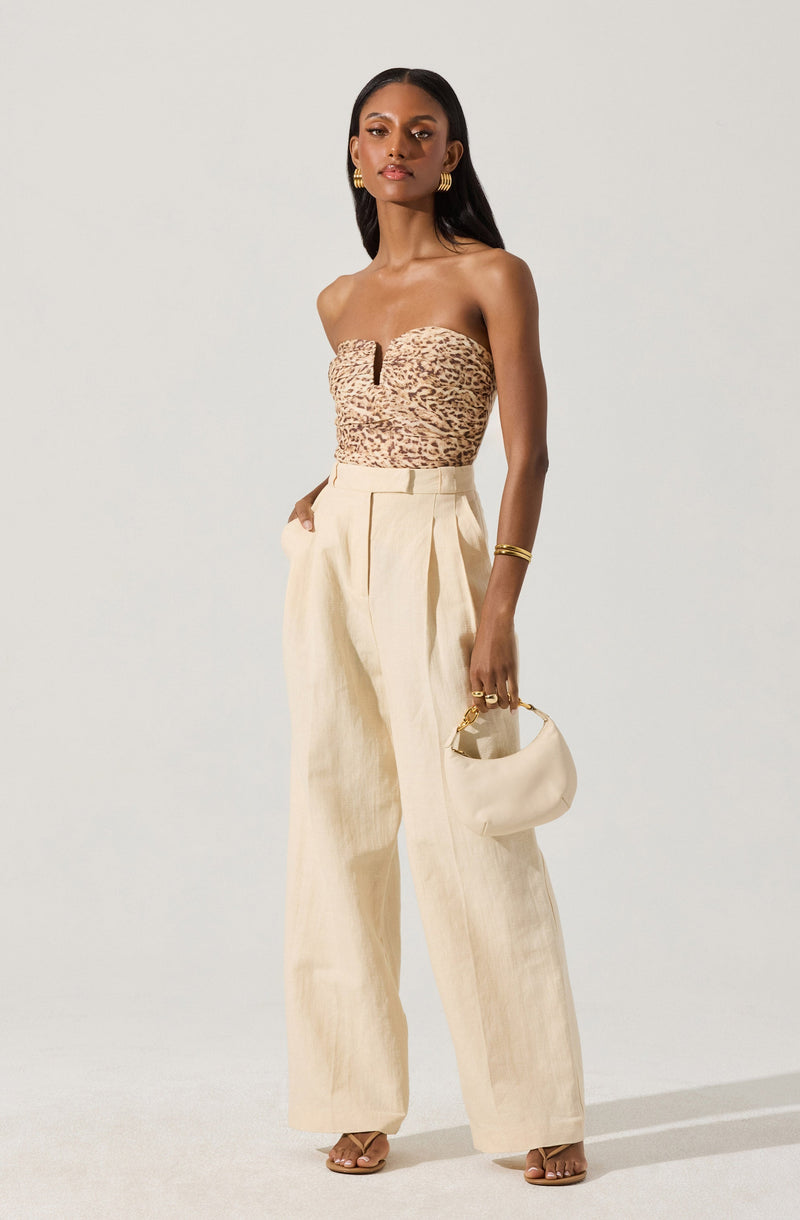 A woman is wearing a strapless, fitted top with a leopard print pattern in shades of brown and beige. She is also dressed in high-waisted, wide-leg beige trousers with pleats at the front. She accessorizes with gold hoop earrings, a gold bracelet, and rings, and carries a small beige purse with a gold handle. She is wearing open-toe beige high-heeled sandals.