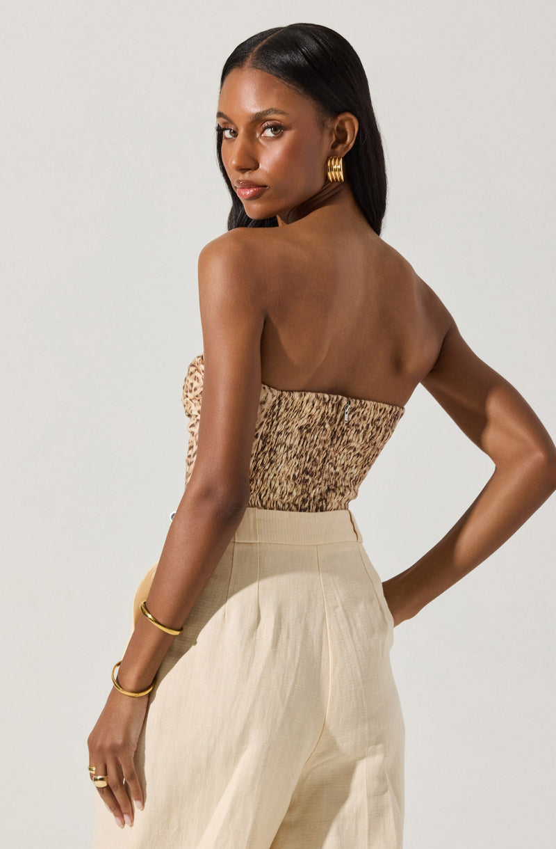 A woman is wearing a strapless, fitted top with a animal print pattern in shades of brown and beige. She pairs it with high-waisted, wide-leg beige pants. She accessorizes with gold hoop earrings, gold bracelets on her left wrist, and a gold ring on her left hand.