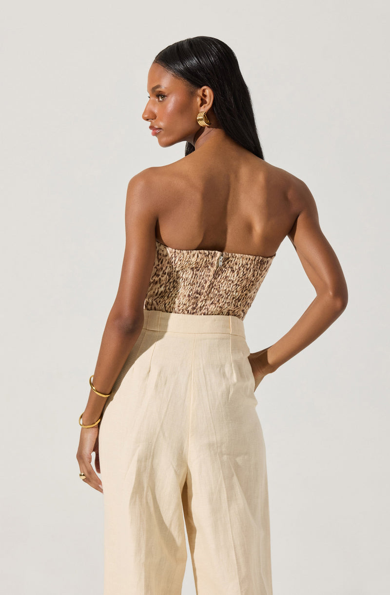 A woman is wearing a strapless top with a textured, animal-print pattern in shades of brown and beige, and high-waisted, cream-colored wide-leg trousers. She has accessorized with gold hoop earrings, a gold bracelet, and rings.