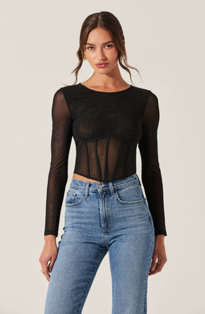 A woman is wearing a black long-sleeve top with sheer, see-through fabric and structured paneling at the waist. She is also wearing high-waisted, light-wash blue jeans.