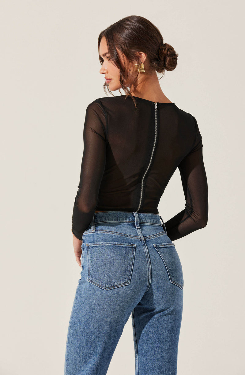 A woman is wearing a sheer black long-sleeve top with a central silver zipper running down the back. She has paired it with high-waisted blue jeans that have a classic five-pocket design. Her dark brown hair is styled in a low, messy bun.